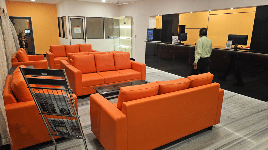 A reception area with a black front desk and a staff member standing behind it, adjacent to a waiting lounge with bright orange sofas - Hotel Pandian