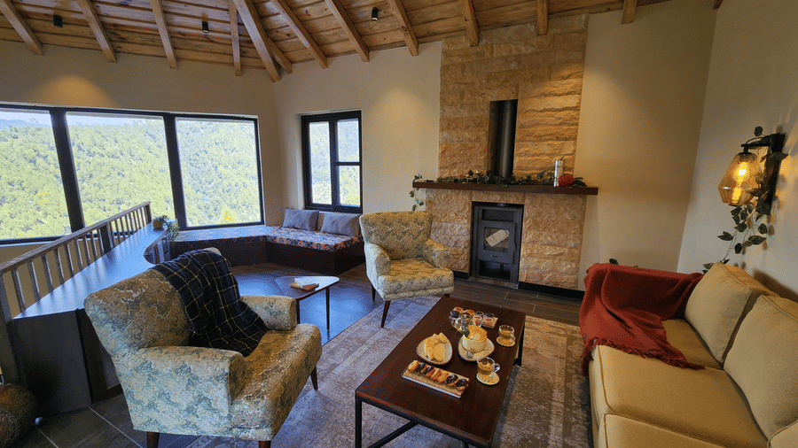 First floor lounge under an exposed timber ceiling, with a stone fireplace, window seat and corner glazing over the wooded valley at The Chamomile Estate.