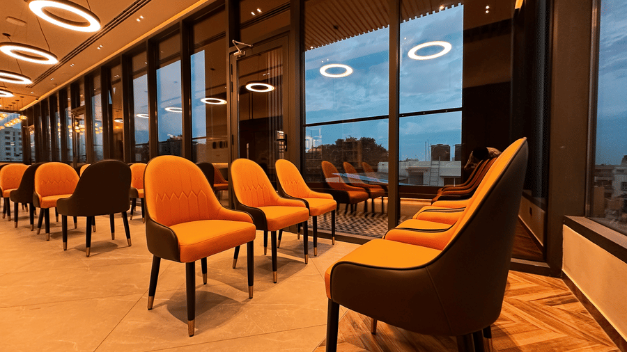 6 orange-coloured cushioned chairs placed in the restaurant facing each other with a huge window partition overlooking the city views at the restaurant at F Hotels, Bengaluru.