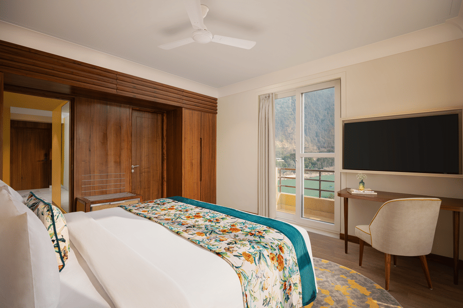 Bright hotel bedroom featuring floral linens, wooden paneling, a desk area, and a balcony with a scenic river view.