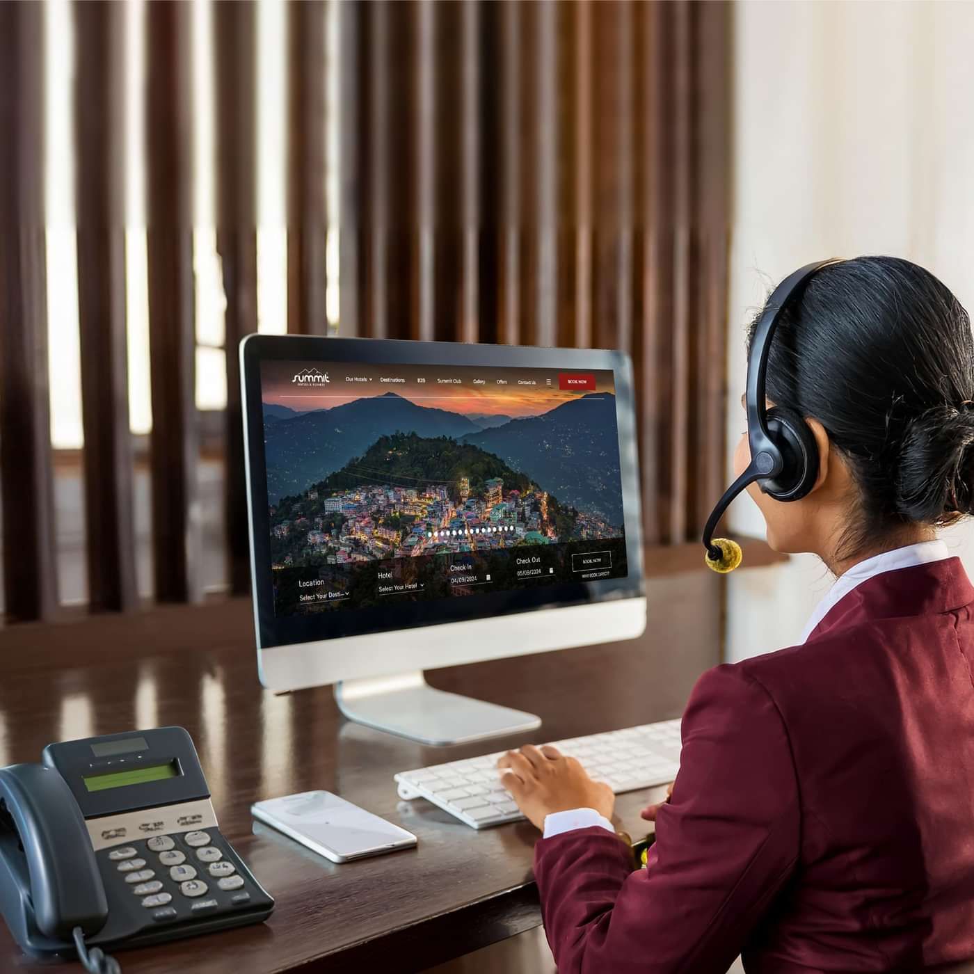 Female customer service representative in uniform using a headset and computer at a desk, assisting with hotel bookings on a website displaying a scenic town.