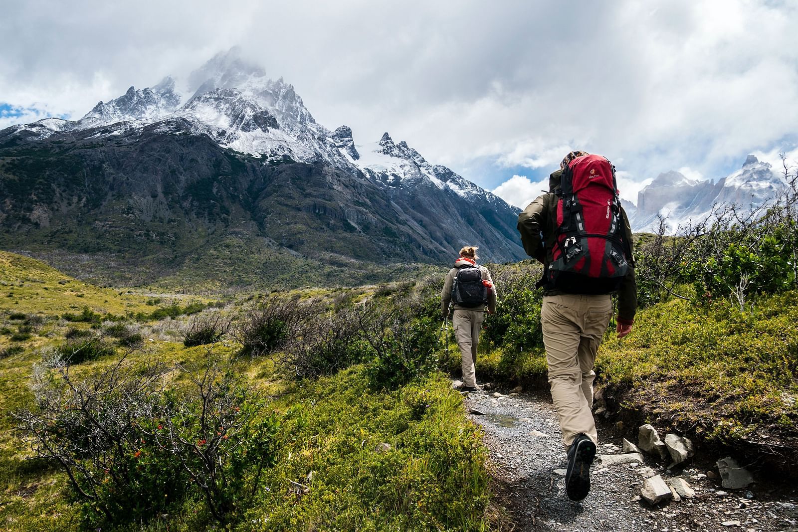 Two people walking on a trekking path with greenery around them and a snow-clad mountain in the background