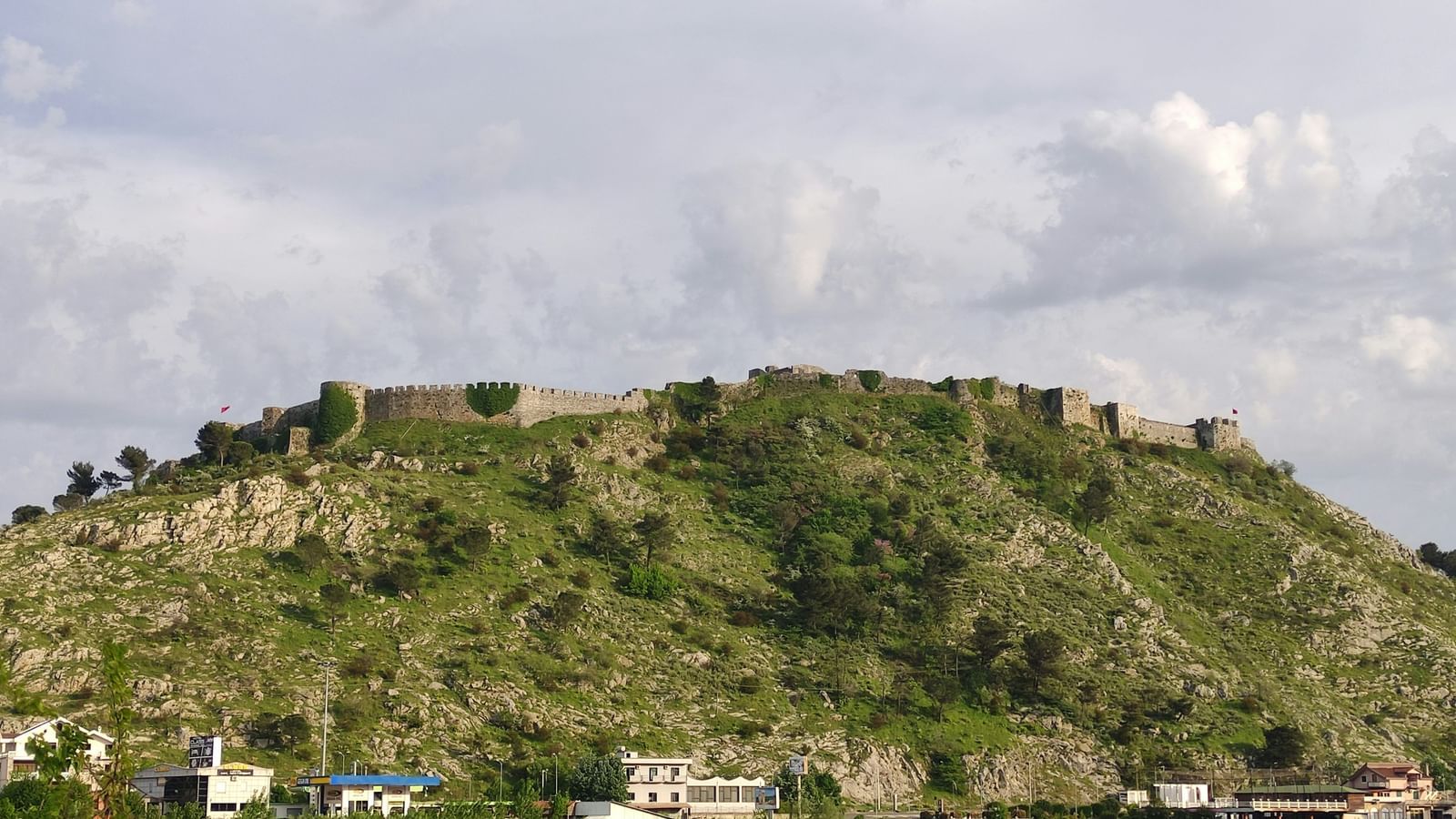 A panoramic shot of a low, rolling green hill range with an ancient stone fortress or structure perched on its peak against a cloudy sky.