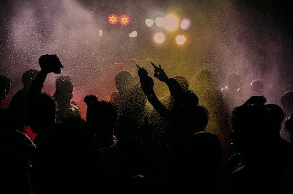  Silhouetted figures with raised hands at a lively, crowded event with bright, colourful stage lighting.