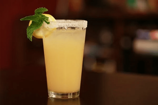 A tall glass filled with a beverage, garnished with a citrus slice and mint leaves, sits on a dark surface with a blurred background.