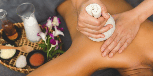 A person is receiving a back massage with an herbal compress poultice while lying down next to a basket of spa ingredients.