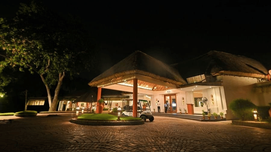 A night exterior view of Vedic Village Spa Resort featuring a warmly lit thatched roof entrance building surrounded by tall trees under a dark evening sky.