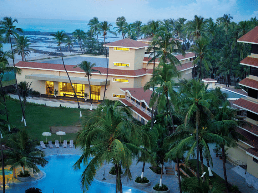 Aerial dusk view of a beachfront resort with warm lit buildings surrounded by tall palm trees and a blue swimming pool in the foreground with the sea visible in the background.