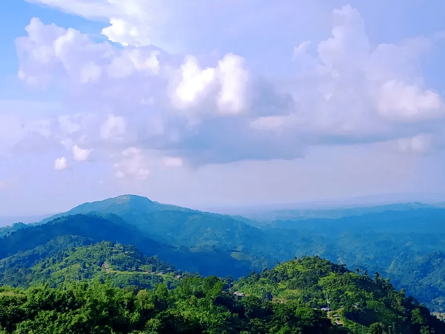 Expansive view of layered hills beneath a bright sky with scattered clouds and rich green forest cover.