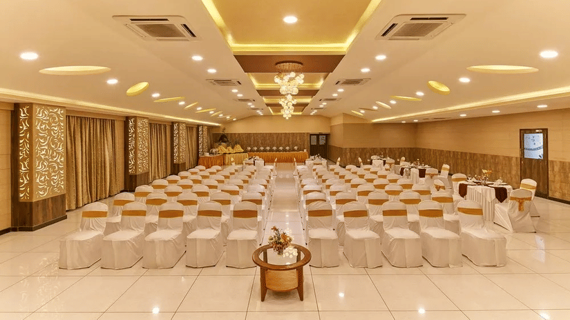 A banquet hall is arranged in theatre-style seating with rows of white-clothed chairs featuring gold sashes, brightly illuminated by a modern chandelier and recessed ceiling lights at Ramanashree Richmond.
