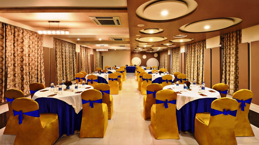 An elegantly decorated banquet hall at Lords Inn Porbandar, with round tables set for dining and chairs with blue and gold accents.