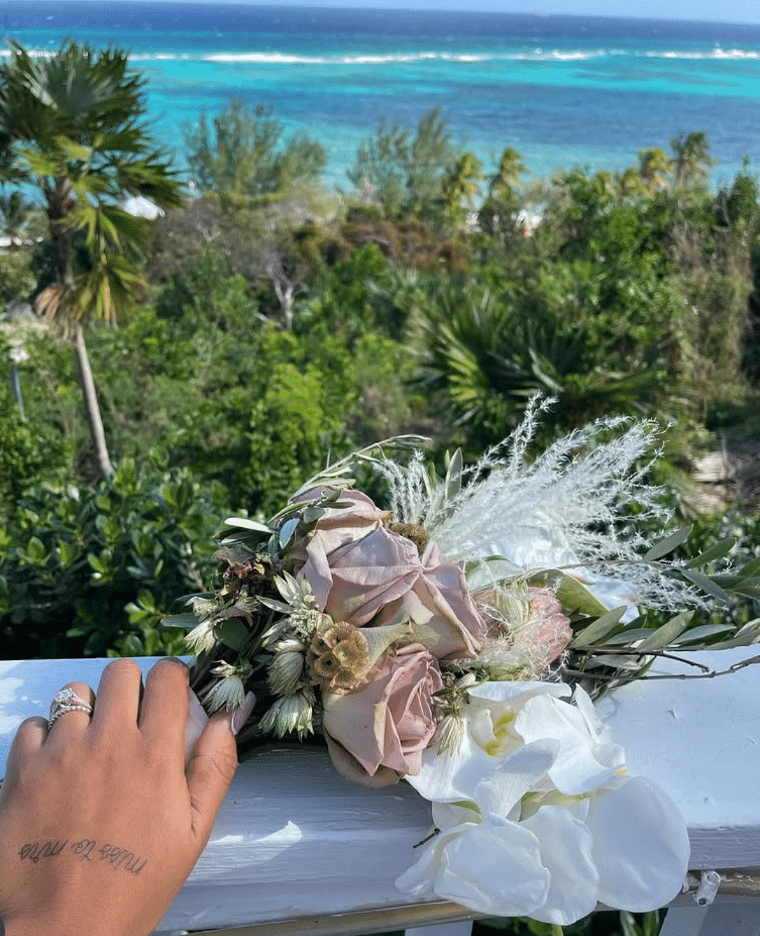 A hand holding a wedding bouquet of roses and orchids with ocean and trees in the background | A Stone's Throw Away 