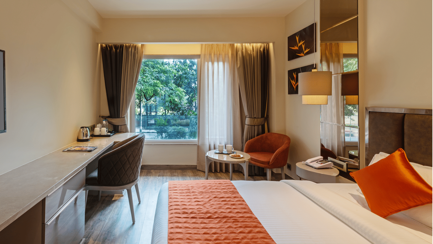 Modern Hotel Saket 27 room with bed, orange runner, armchair, work desk, and large window with greenery view.