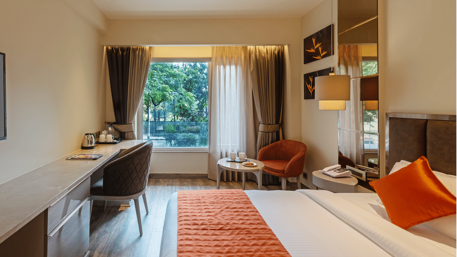 Modern Hotel Saket 27 room with bed, orange runner, armchair, work desk, and large window with greenery view.