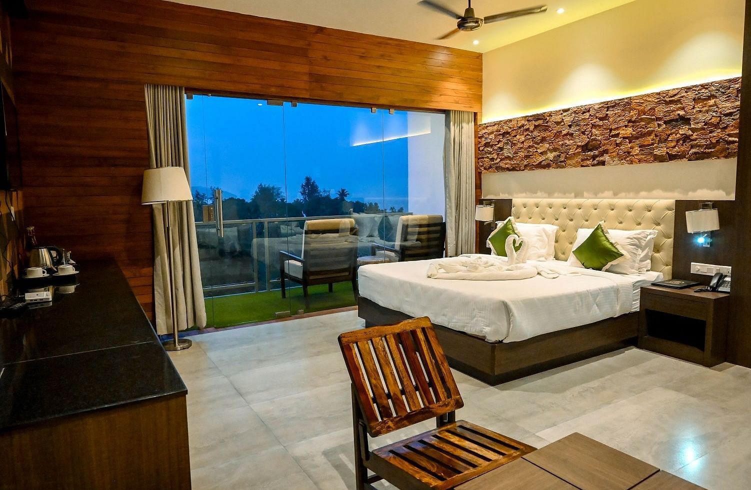 A hotel bedroom with a large bed, a comfortable armchair, and a wide view of the outdoors through a large glass door leading to a balcony - Sea Hills Hotels, Sri Vijaya Puram (Port Blair)