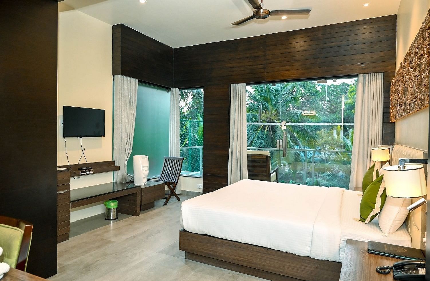 A hotel room with a large bed, a television, and a panoramic window revealing a vibrant green landscape outside, under a ceiling fan - Sea Hills Hotels, Sri Vijaya Puram (Port Blair)