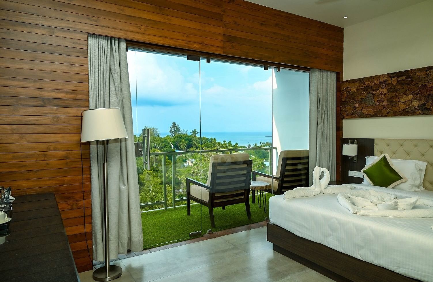 A hotel room interior featuring a large bed, a bedside lamp, and a full-length glass door leading to a balcony with an outdoor seating area, all against a backdrop of wooden wall panelling - Sea Hills Hotels, Sri Vijaya Puram (Port Blair)
