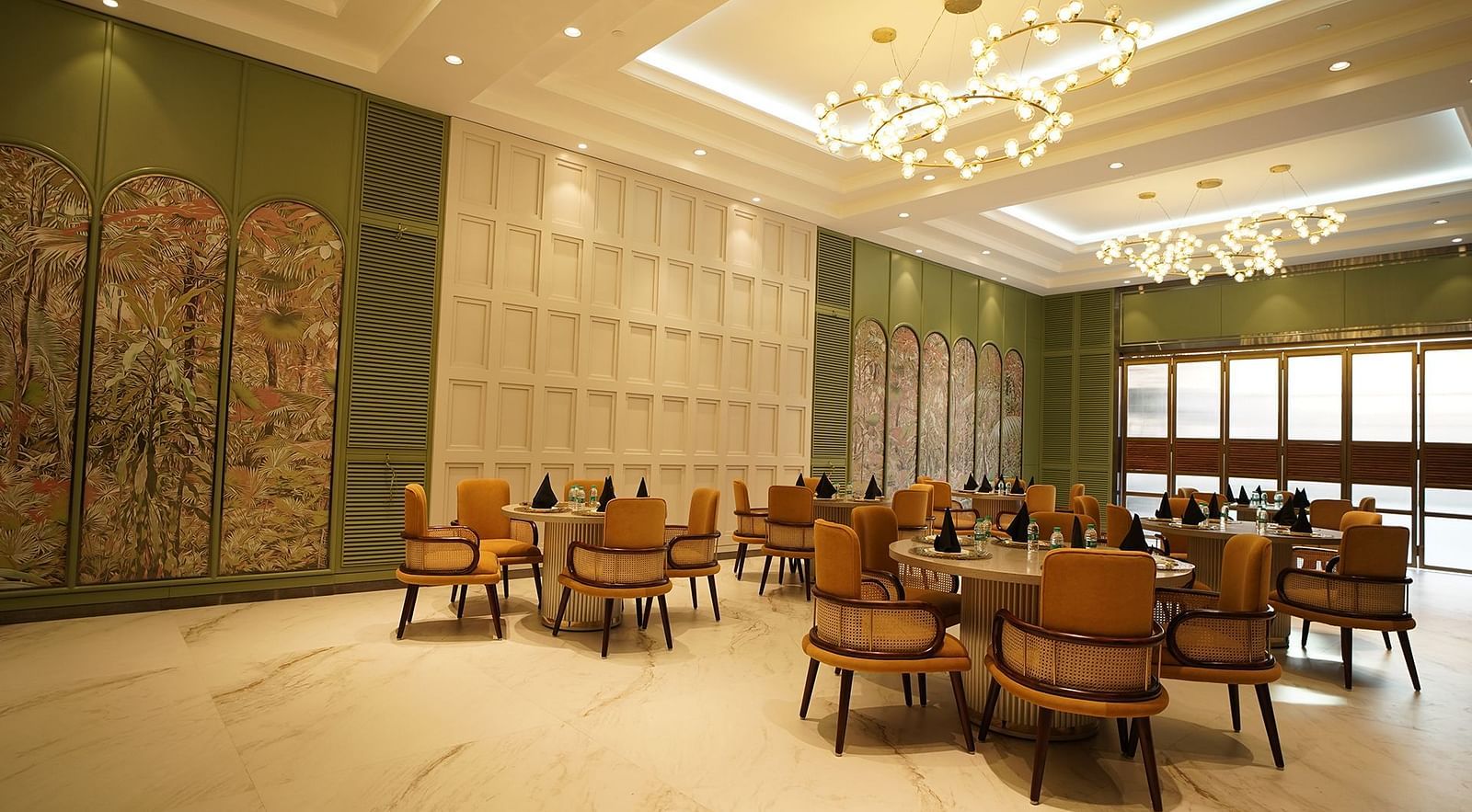  A stylish dining area at Nirvana Luxury Hotel, Ludhiana, featuring round tables with tan chairs, modern chandeliers, and elegant wall decor.