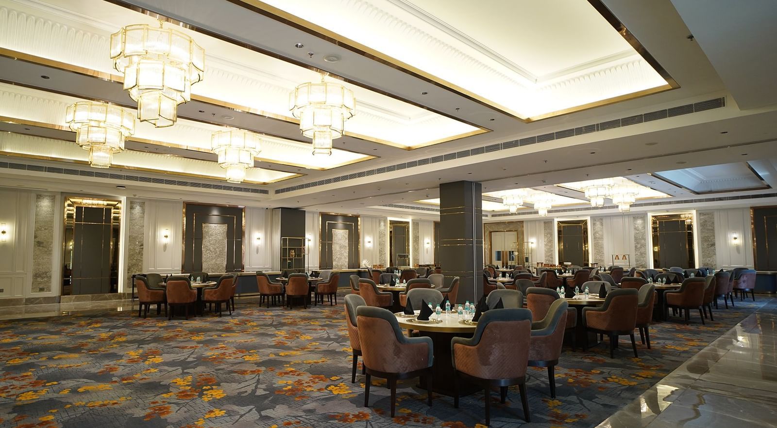 An elegant banquet hall at Nirvana Luxury Hotel, Ludhiana, featuring round tables with plush chairs on a patterned carpet, illuminated by multiple tiered chandeliers and recessed lighting.