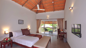 A spacious bedroom with twin beds, soft lighting, a wooden bench, and a large glass door leading to a balcony with nature views.- Abad Brookside Lakkidi, Wayanad