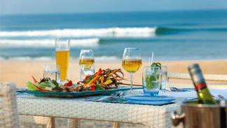 A delightful beachfront dining setup with a variety of drinks and a plate of fresh seafood, perfect for enjoying a meal by the ocean.