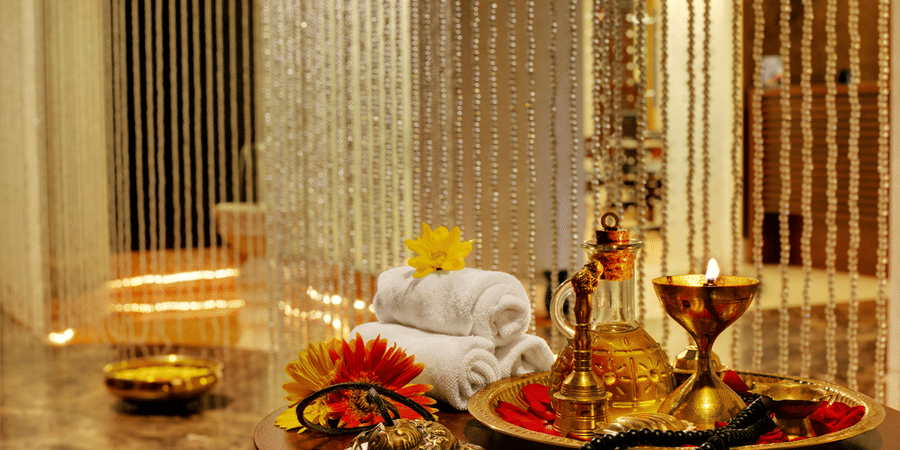 Myra Spa at Kenilworth Resort & Spa, Goa, featuring rolled white towels, a brass tray with massage oil, a lit lamp, fresh flowers on a round wooden table, and shimmering beaded curtains in the background.
