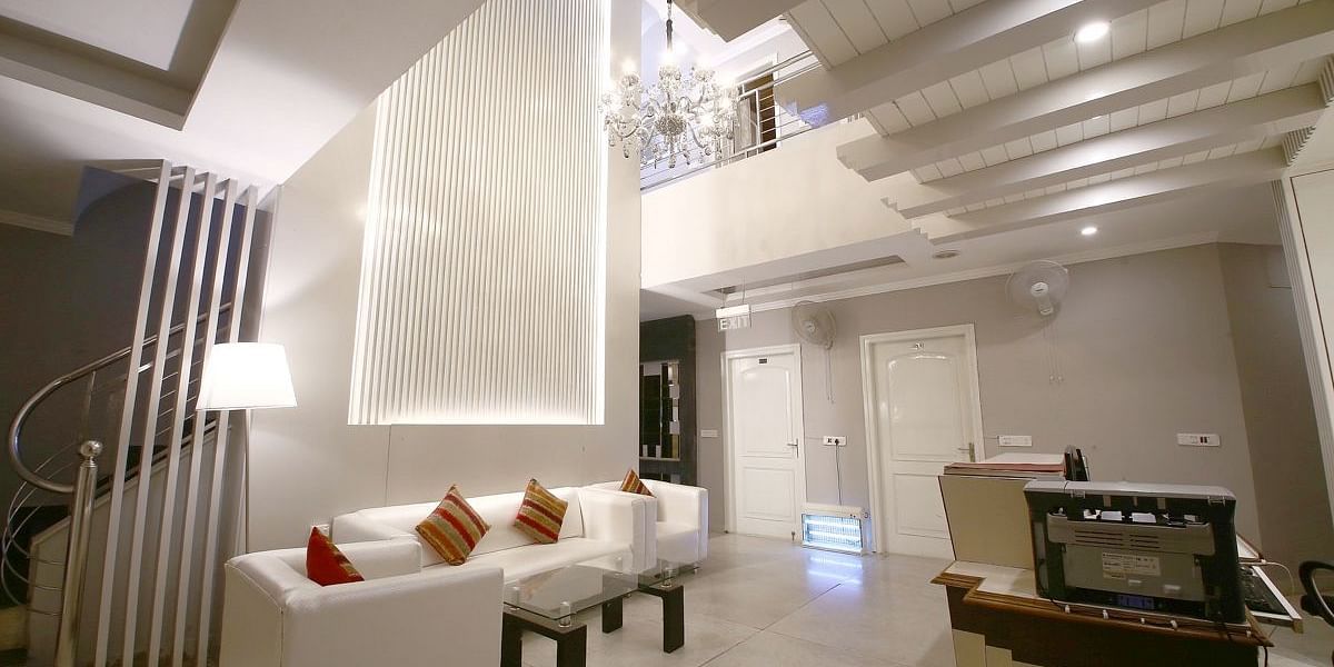 A hotel lobby with a white sofa, decorative pillows, a textured wall with lighting, a spiral staircase, and a reception desk, lit by a chandelier. - Hotel Diamond Plaza, Chandigarh.