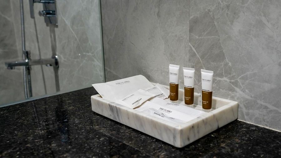 A close-up of a bathroom countertop with a white marble tray holding three small tubes of toiletries and a neatly folded paper cover. The background features grey marble wall tiles and a chrome shower fixture. - Clarks Inn Suites, Near Taj Mahal