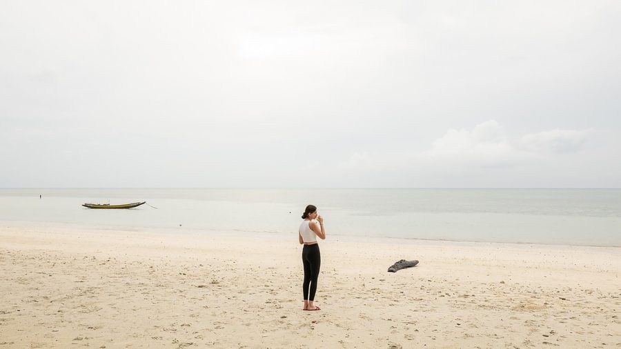 Woman standing on a wide, empty beach looking at the calm ocean under a pale sky.