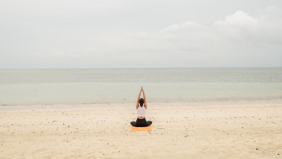 Woman practicing yoga in a seated pose on an orange mat on a beach facing the sea.