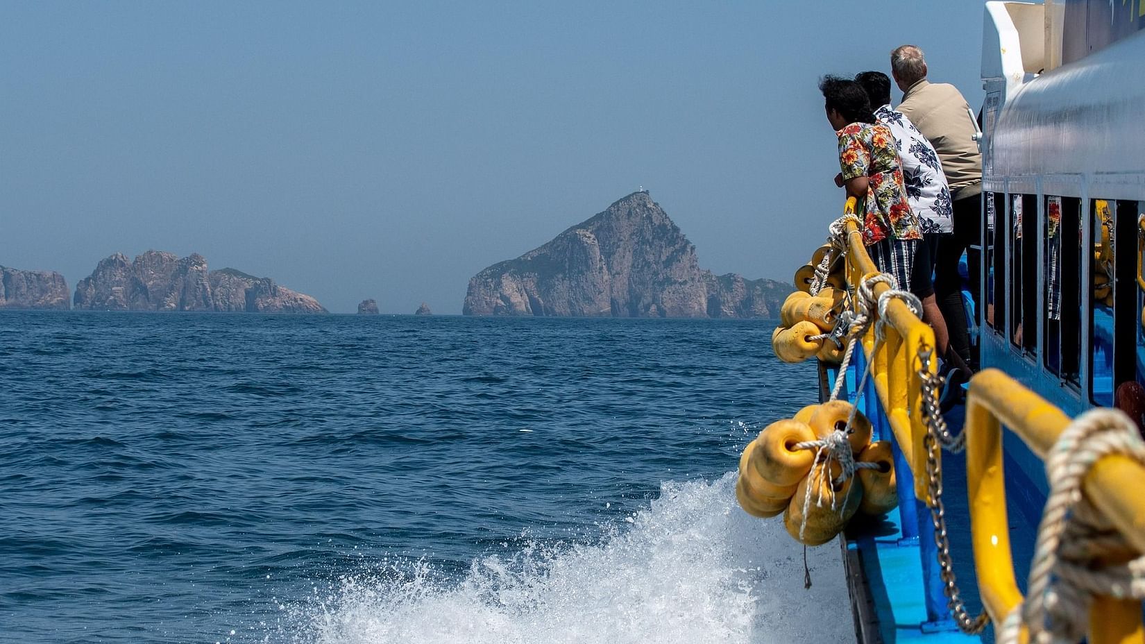 People stand on the deck of a boat moving through the blue sea, with rocky islands visible in the distance under a clear, pale sky.