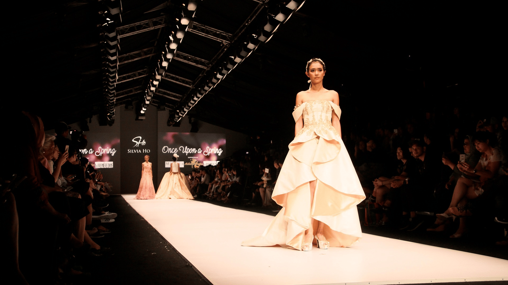 A model walks a long, lit runway in a structured cream couture gown with a high-low hemline under dark studio lights.