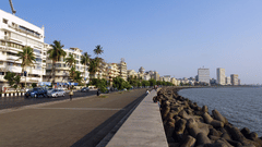 A wide promenade with people walking along a seafront lined with buildings and palm trees.
