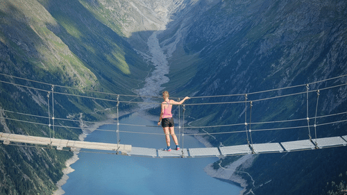 A lady standing on a bridge surrounded by mountains and a water body below