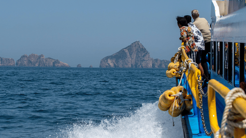 People stand on the deck of a boat moving through the blue sea, with rocky islands visible in the distance under a clear, pale sky.