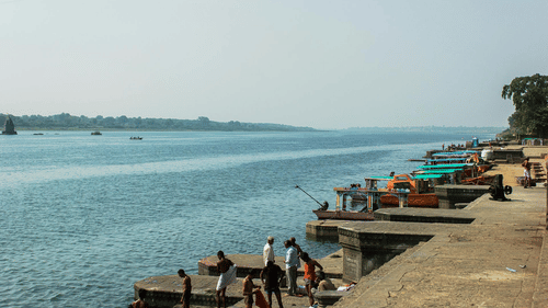 People gathered along a riverbank with boats docked, engaging in various activities on a sunny day.