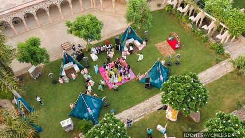 An aerial view shows an event with blue tents and people gathered on the lawn of Tijara Fort-Palace - 19th Century, Alwar, surrounded by trees and historic walls.