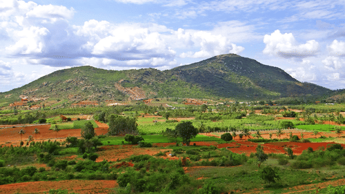 Lush green hills rising above a patchwork of rural farmland under a bright, cloudy blue sky.