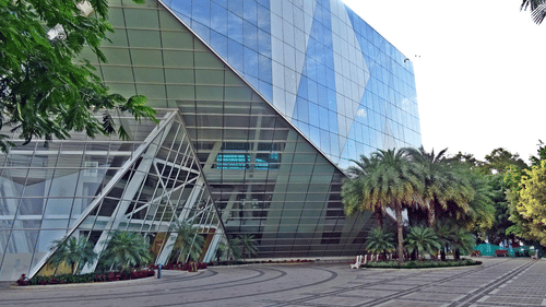 A large, modern glass office building with sharp angles, surrounded by green palm trees and a paved area.