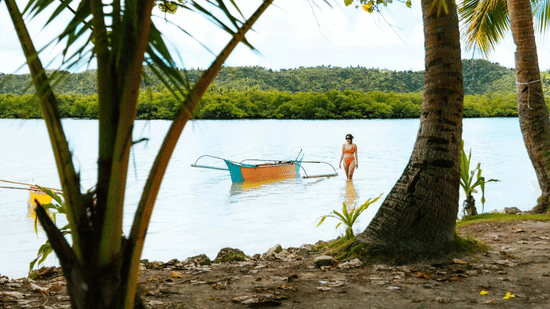 A small colourful boat and a person standing in shallow water framed by palm trees at Lamari Siargao.