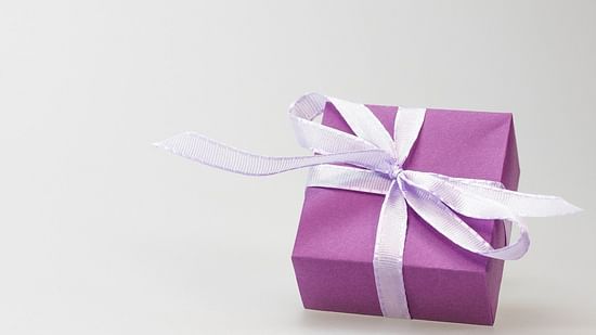 A small, square gift wrapped in purple paper and tied with a white ribbon sits on a plain white surface