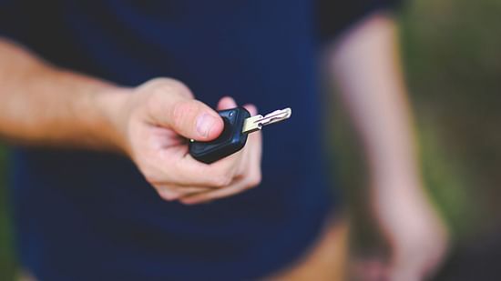 A person's hand holds a car key with a black fob and a metal key blade, against a blurred background of clothing.
