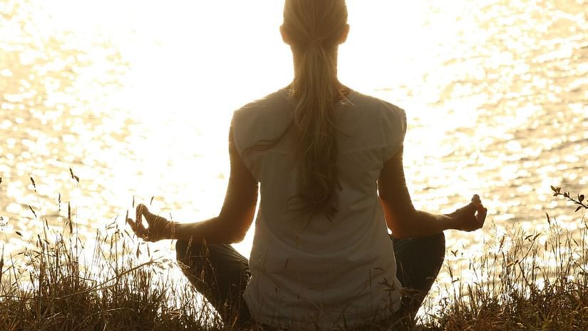 A woman sitting facing the sea and meditating with the sun's reflection shining on the water