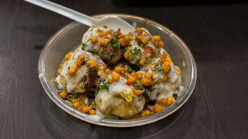 A plate of dahi puri topped with yogurt, sev, chutneys, and coriander, served with a plastic spoon on a dark table.