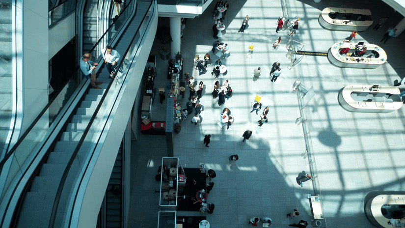 A top-down view of a convention centre with an escalator on the left hand side, and people near the registration desk and other showing areas at ITB India 2026.