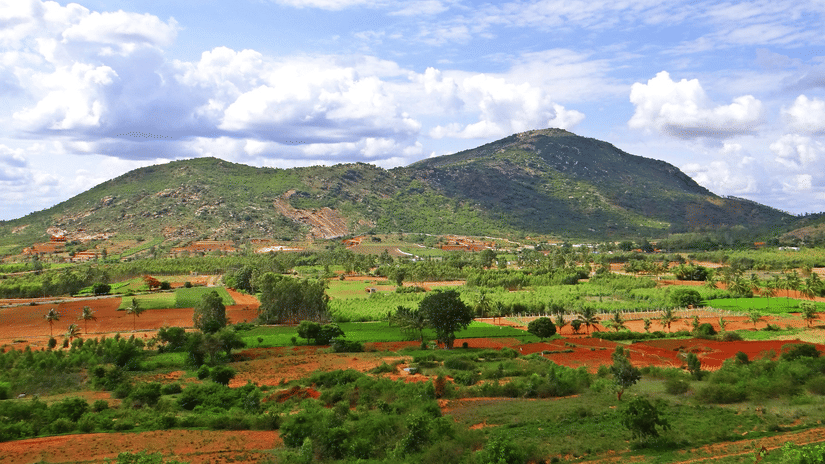 Lush green hills rising above a patchwork of rural farmland under a bright, cloudy blue sky.