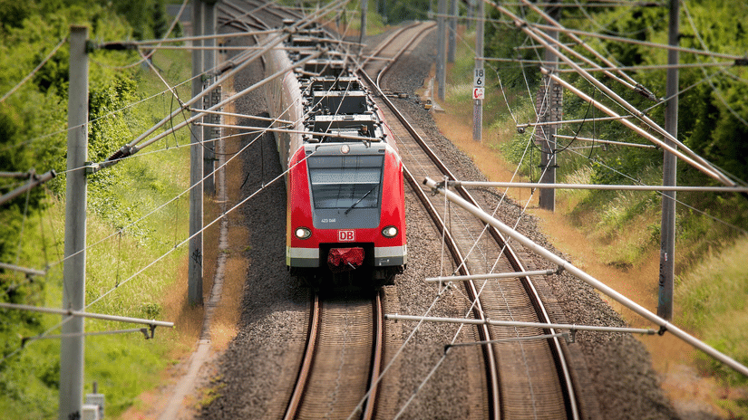 An overhead, slightly blurred shot of a red regional train traveling along a curved track through a lush, green landscape under bright daylight.