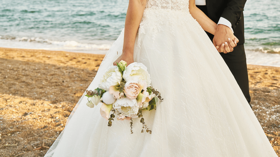 alt-text Bride in white wedding gown holding hands with groom at a beach