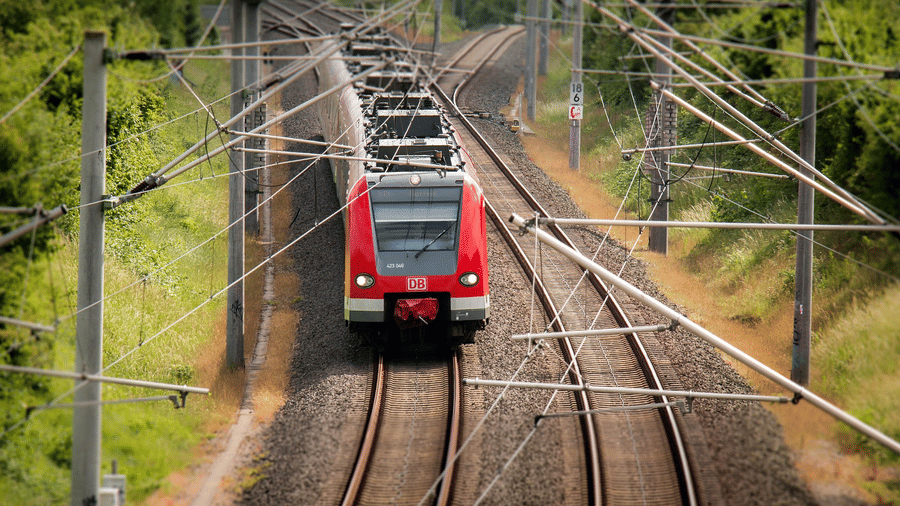 A red Deutsche Bahn passenger train travelling along railway tracks lined with overhead power lines, surrounded by green trees and shrubs.
