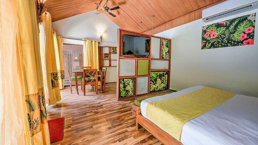 Spacious resort room with a king-size bed, wooden partition with built-in TV and greenery-themed artwork, dining area with wooden chairs, and large windows displaying pool views.